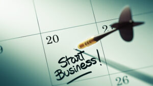 A close-up of a calendar page with a date marked on the 20th. A black and gold dart is pinned directly into the square for the 20th, which has the handwritten words, 'Start Business!' written in bold black ink with an underline and exclamation mark. This image conveys a sense of goal-setting for launching a business.