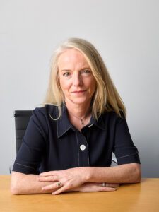 Professional portrait of Catherine Stack, she has long blonde hair and is waring a navy blue short-sleeved blouse with white stitching. She looks calmly at the camera and has her arms gently folded on the table in front of her. She is sitting in an office chair.