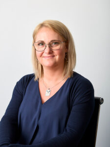 Professional headshot of Paulina Klinger, she has straight, shoulder-length blonde hair and glasses. She smiles gently, she is wearing a dark navy blouse with a V-neckline and a distinctive silver rectangular pendant necklace. She has subtle gold circular drop earrings. She sits in an office chair.