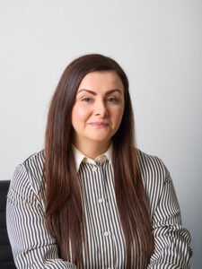 Professional headshot of Rosarii Doyle, she has long, straight brown hair. She is wearing a vertically striped black-and-white button-down shirt with a collar. She is smiling gently, and she sits in an office chair.