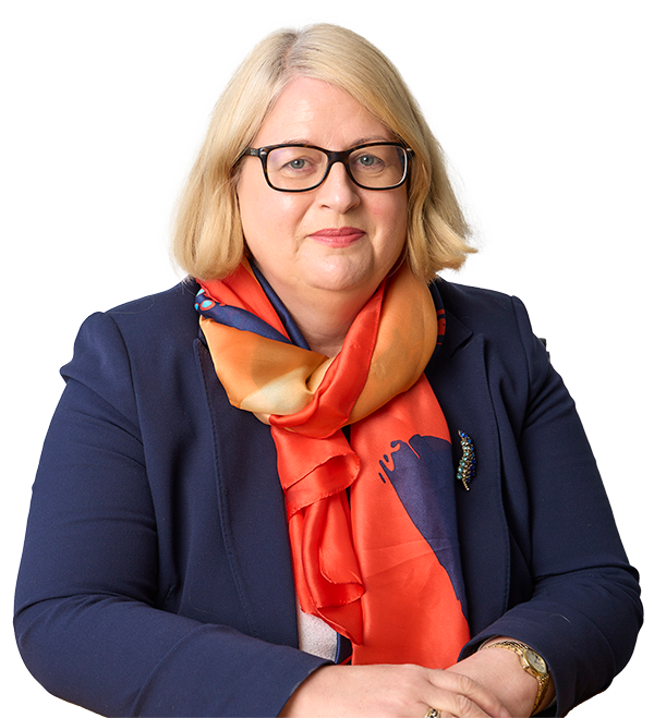 Professional headshot of Sarah Breslin, she has shoulder-length blonde hair and glasses with a black frame. She is wearing a navy blue blazer adorned with a decorative brooch and a vibrant silk scarf in shades of orange, red and blue. Her hands are gently folded in front of her and she wears a gold wristwatch and ring.