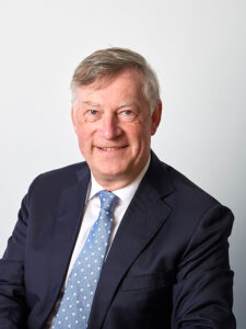 Professional headshot of Val Stone, he has short, grey hair and he is wearing a dark navy suit, a white shirt and a blue tie with a white polka dot feature. He is smiling warmly at the camera.