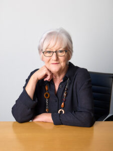 Professional headshot of Veronica Culleton, she has short white hair, and is wearing glasses and a dark shirt with a chunky brown and orange beaded necklace. She is resting her chin thoughtfully on her hand and smiling gently. She is resting her arms on the desk in front of her.